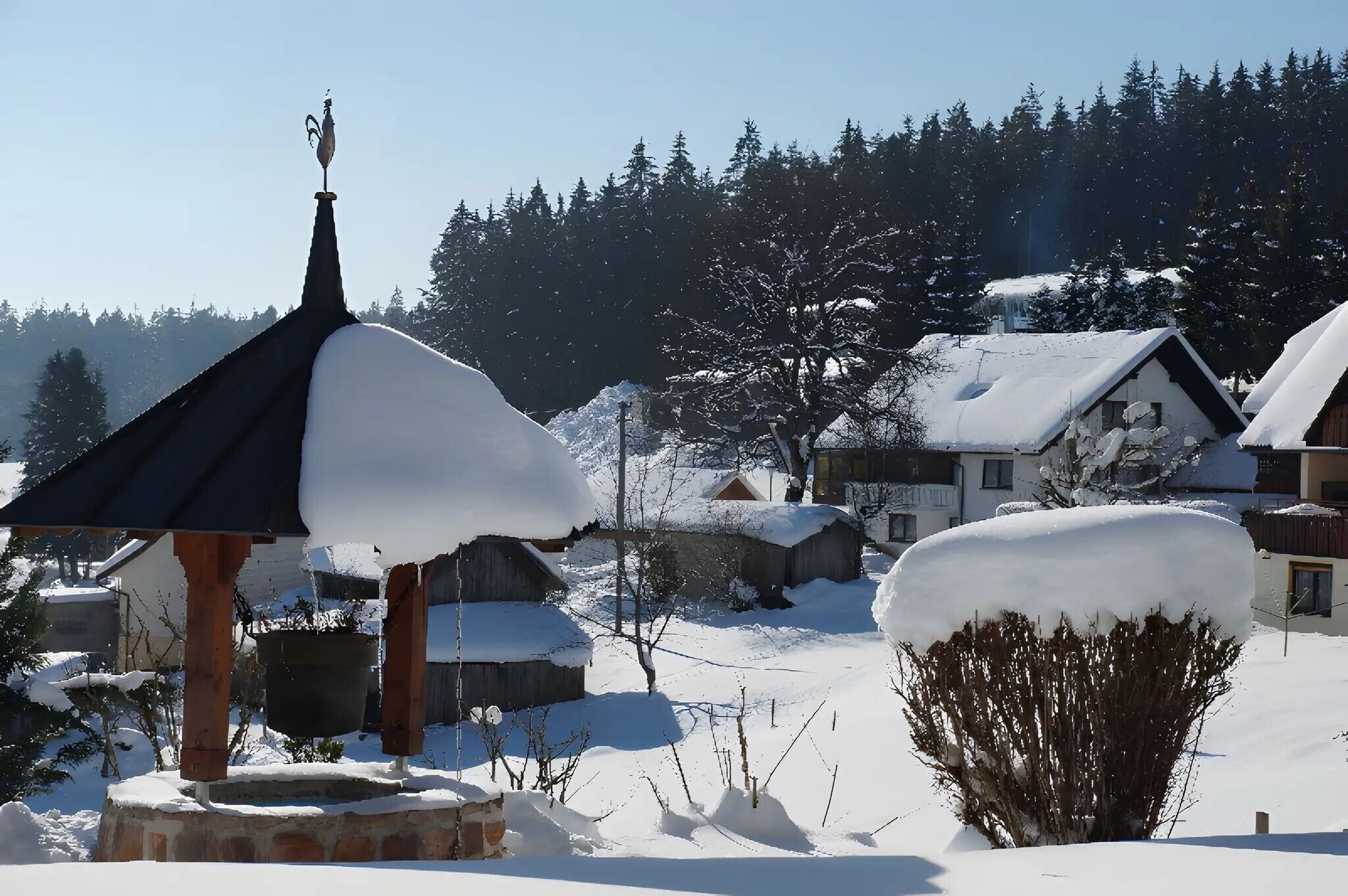 A snow-covered village with traditional houses, a wooden gazebo, and a clear blue sky.
