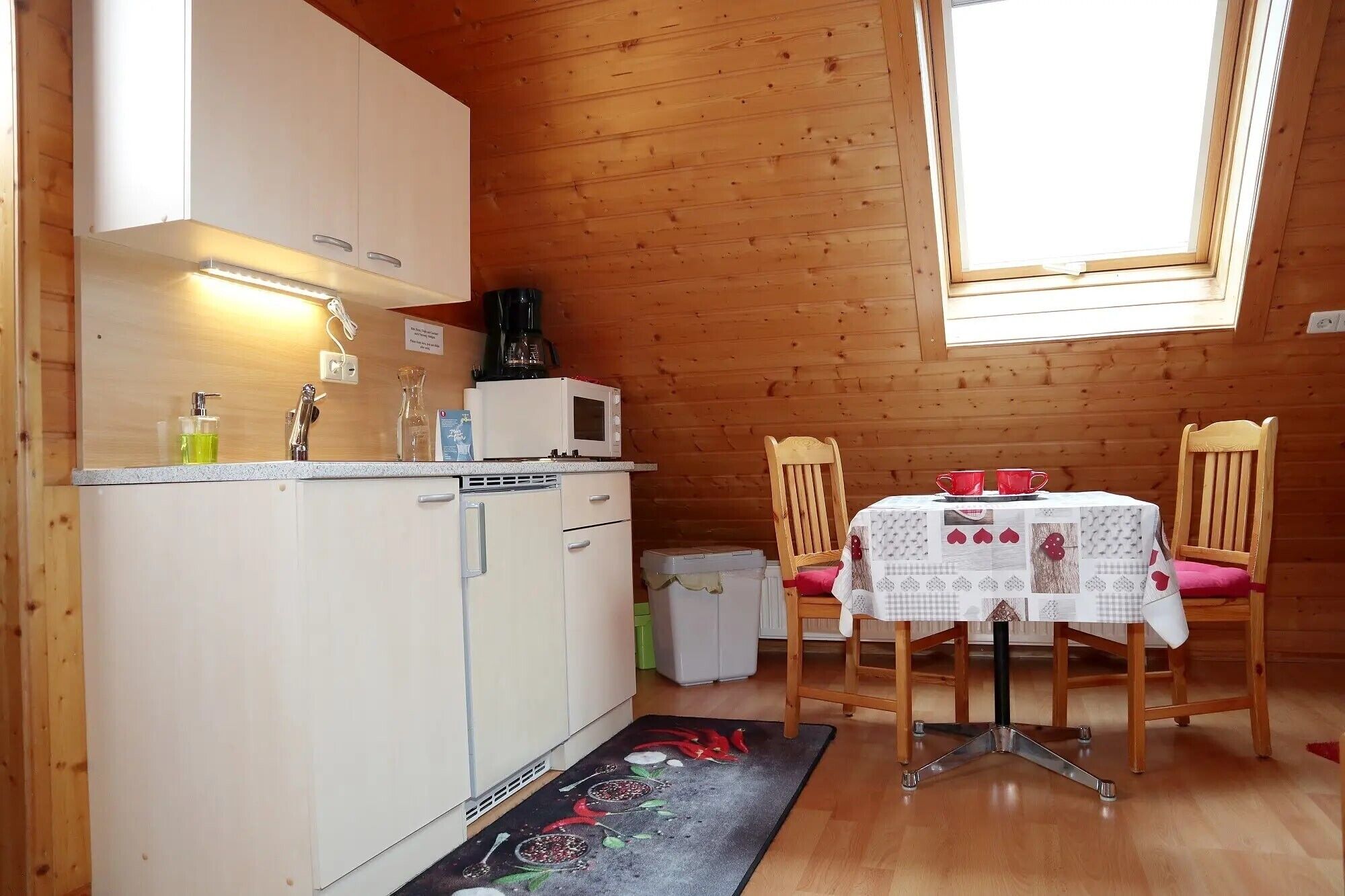 A compact kitchen with a microwave, coffee maker, and a small sink. There's a dining table with chairs and a window above it.