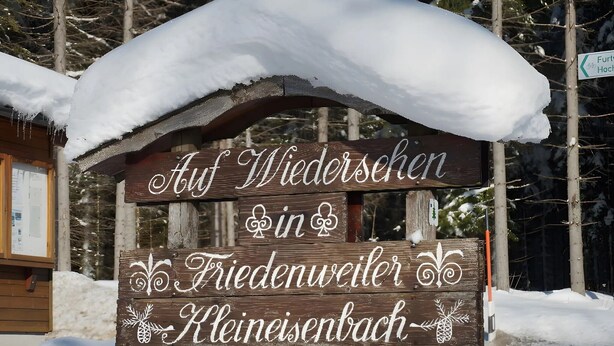 Un letrero de madera con texto en alemán, parcialmente cubierto de nieve, que indica un lugar en Kleinisenbach.