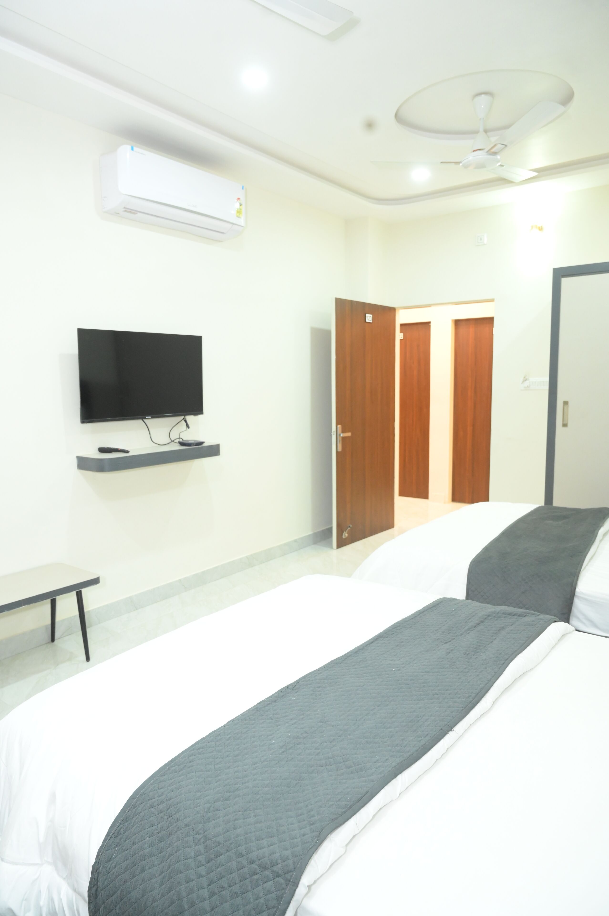 Habitación de hotel con cama, televisor sobre un soporte, ventilador de techo y puerta que da a otra habitación.