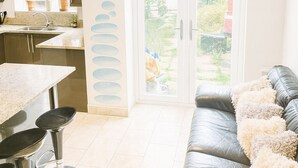 A modern kitchen with a dining area, a sofa, and a view of the garden through a glass door. - Room-charming Guest Room Next to Excel c (London)