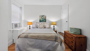 Une chambre à coucher avec un lit, deux lampes de chevet, un tableau au mur, une commode et une fenêtre avec des stores.