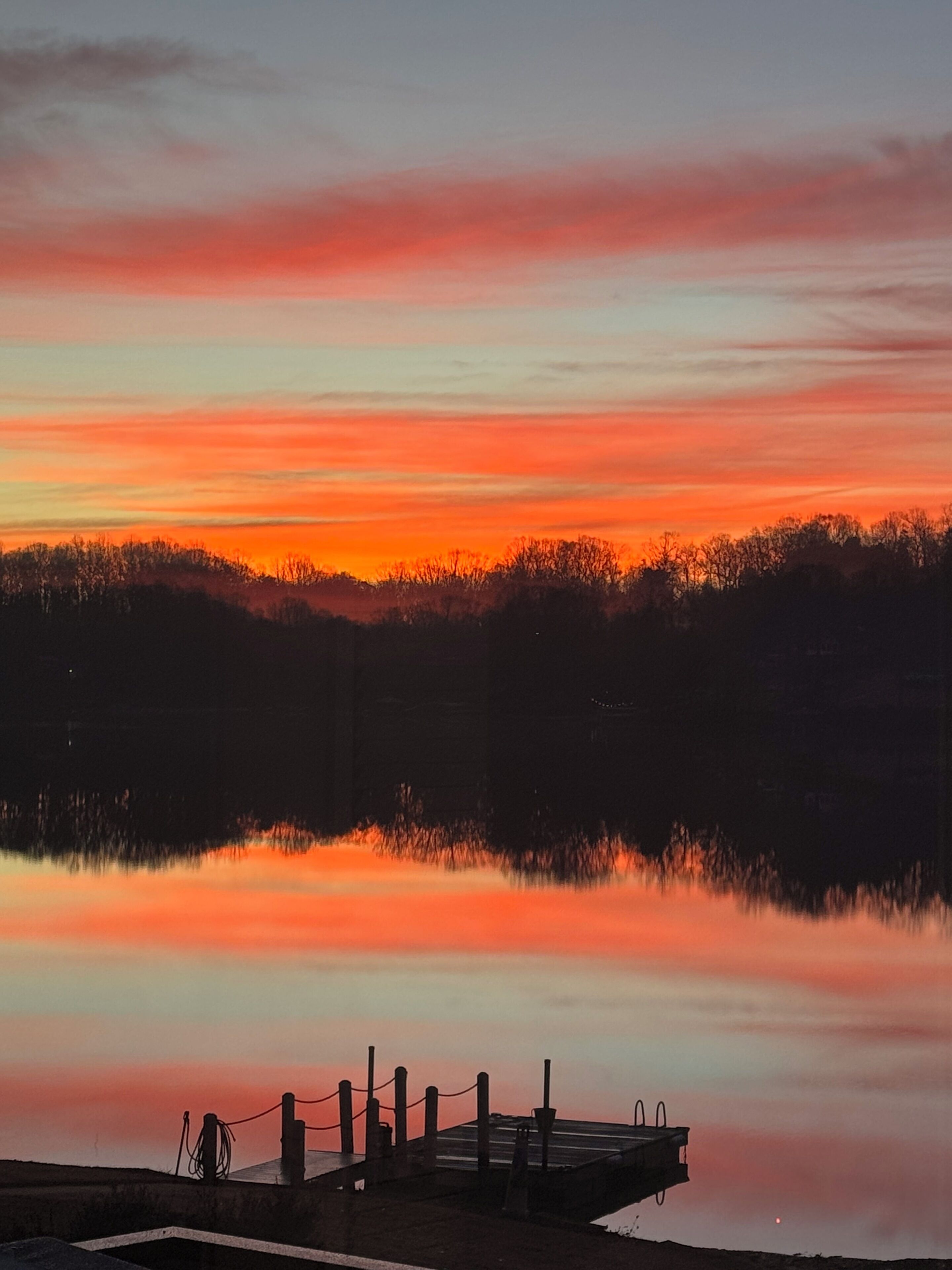 Ein ruhiger See bei Sonnenuntergang mit einem Steg und Bäumen im Hintergrund.