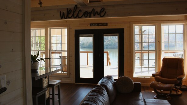 A cabin interior with a leather sofa, a chair, a welcome sign, and a view of a lake through large windows.