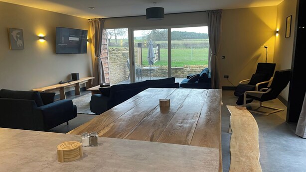 Ein modernes Wohnzimmer mit einem großen Holztisch, schwarzen Sofas und Blick auf einen Garten durch große Fenster.