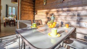 A dining area with a glass table, chairs, and a wooden wall. There is a window with curtains, a table with a floral tablecloth, and a bowl of fruit on the table. - Birds II - Domaine de la Navicelle (Le Pradet)