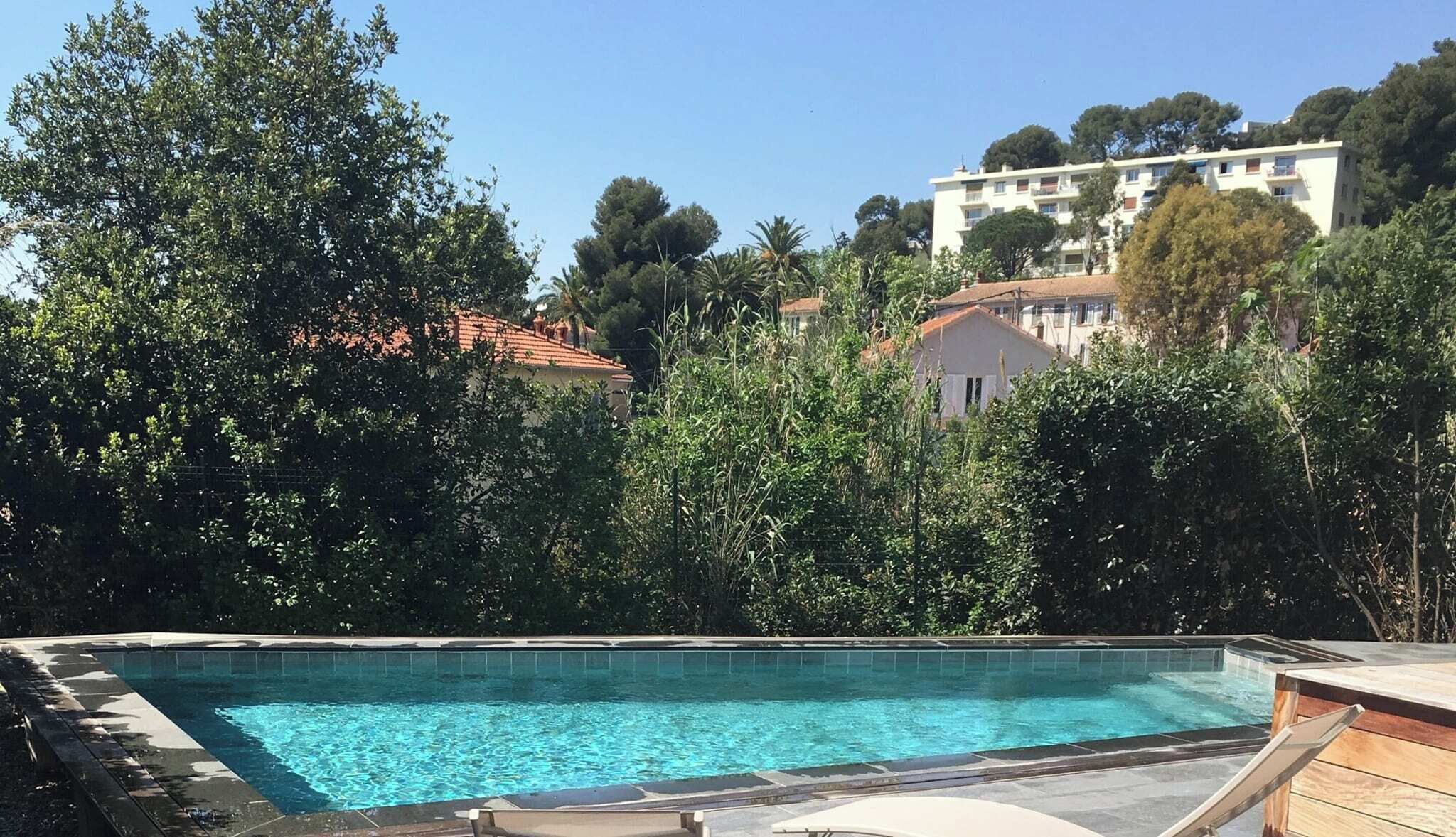Une piscine dotée d’une terrasse en bois et de chaises longues, entourée d’une végétation luxuriante et avec un bâtiment visible au loin.