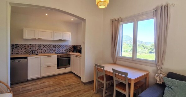 A compact kitchen with white cabinets, a dishwasher, and a stove. There's a dining table with chairs and a window with a view of greenery.