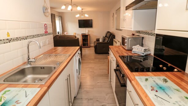 A compact kitchen with a sink, microwave, and stove. There is a sofa and a TV in the background. - Primrose Cottage (Chathill)