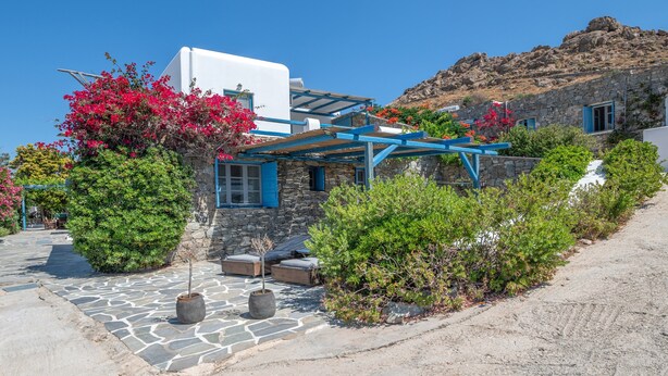 Un edificio in pietra con una parte superiore bianca, un tendone blu e un vialetto in pietra che conduce a una zona con tavoli e sedie.