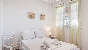 Habitación de hotel limpia y blanca, con una cama, una silla, una mesita de noche con lámpara y dos cuadros en la pared.