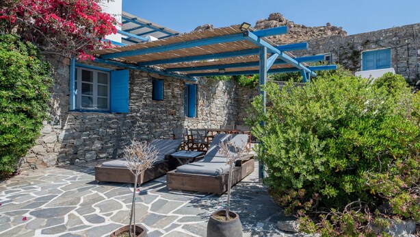 Un edificio de piedra con una pérgola de madera, asientos al aire libre y plantas en macetas.