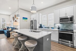 A modern kitchen with a central island, stainless steel appliances, and a dining area with a blue sofa. - Azure on 30A 1-205- A Wave From it All (Santa Rosa Beach)