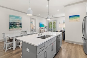 A modern kitchen with a central island, stainless steel appliances, and a dining area with white furniture. - Azure on 30A 1-205- A Wave From it All (Santa Rosa Beach)