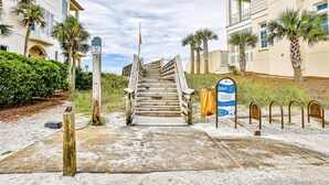 A wooden staircase leading to a building with a balcony, surrounded by palm trees and a sandy path. - Azure on 30A 1-205- A Wave From it All (Santa Rosa Beach)