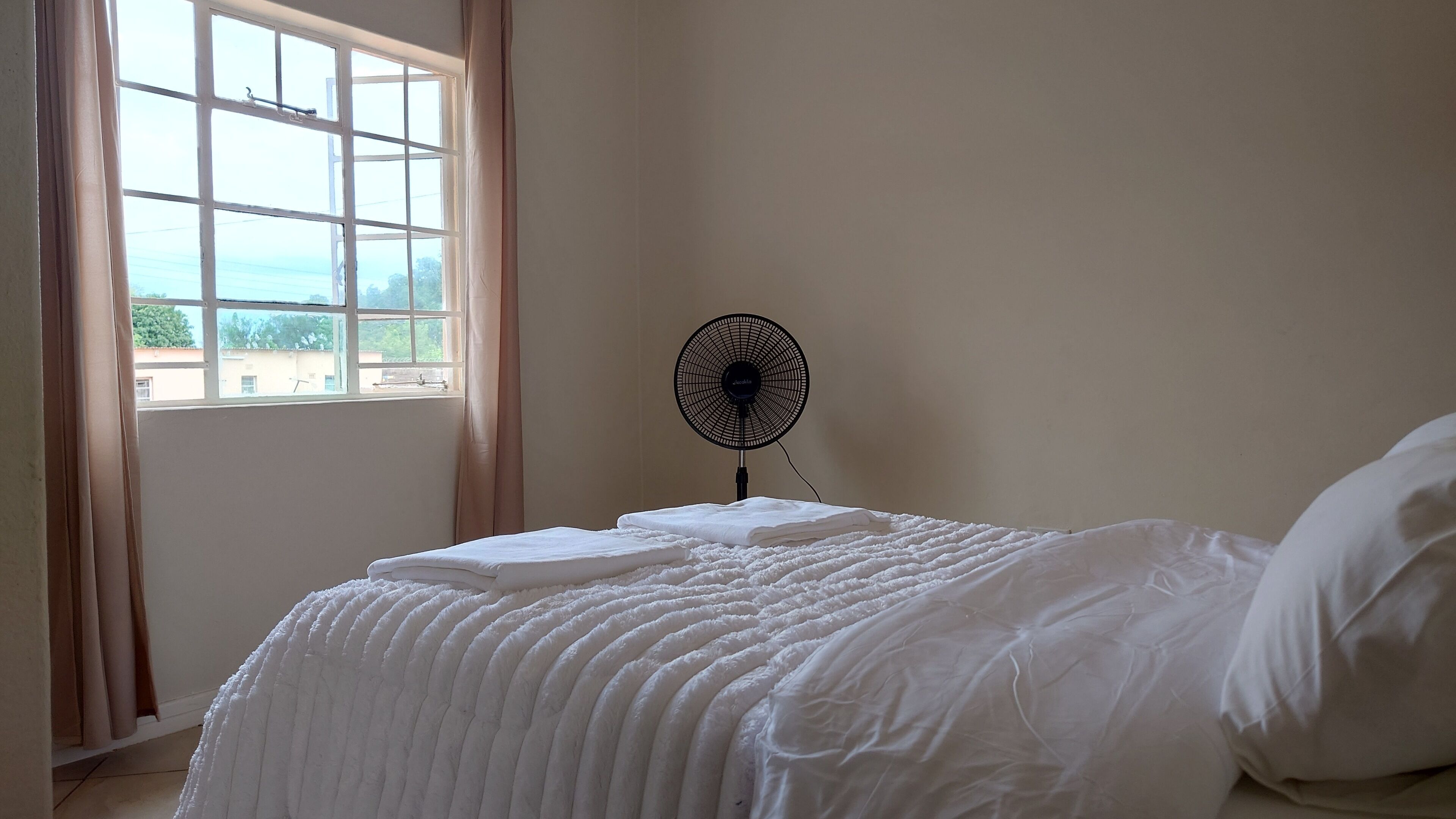 A bedroom with a bed, a standing fan, and a window with curtains.