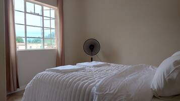 A bedroom with a bed, a standing fan, and a window with curtains.