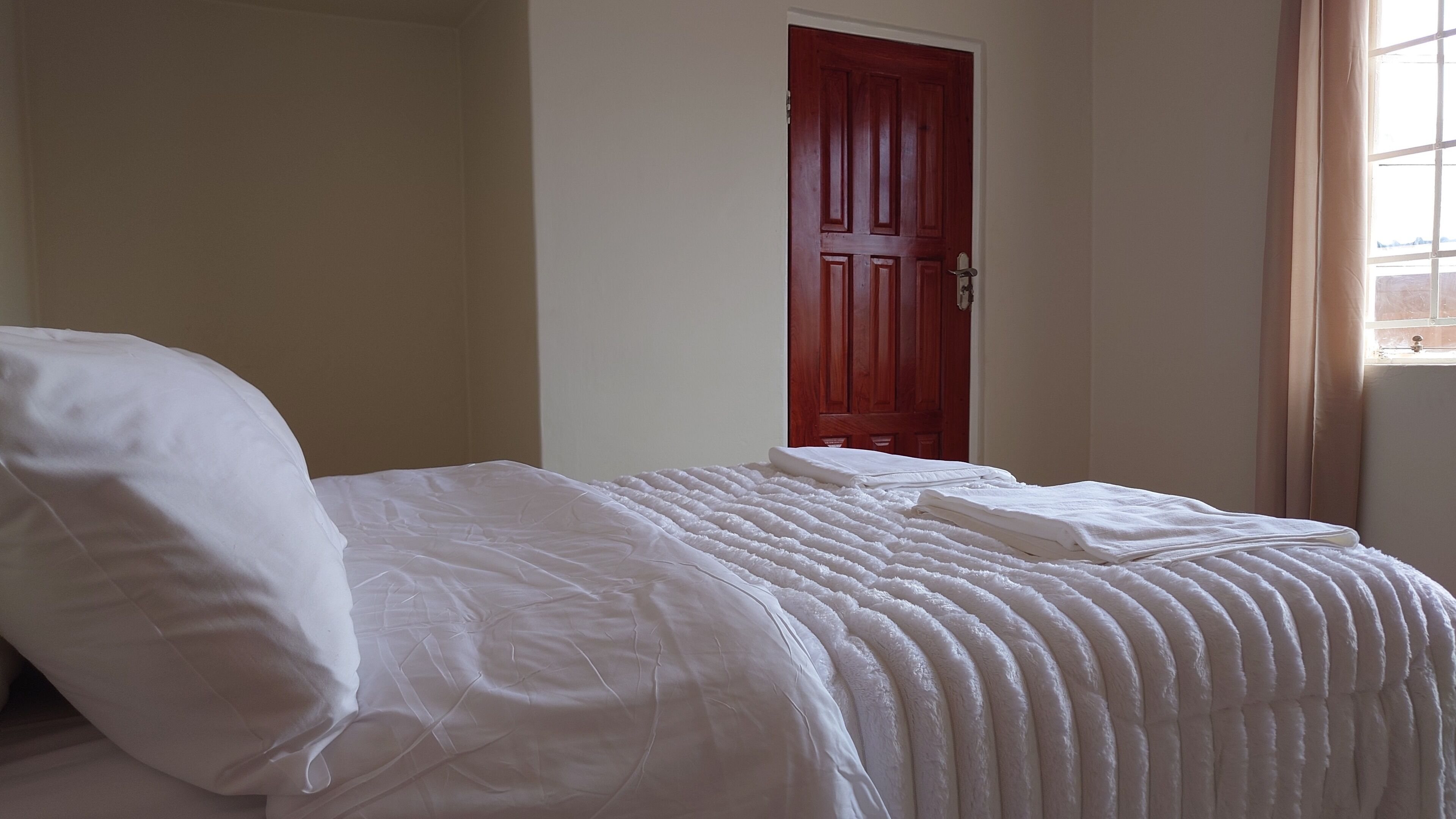 Un letto rifatto con lenzuola bianche in una stanza con una porta rossa e una finestra con tende.