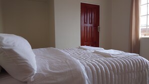A neatly made bed with white linens in a room with a red door and a window with curtains.