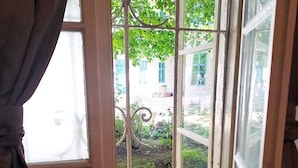 Une pièce dotée d’une grande fenêtre, de rideaux et offrant une vue sur un jardin et un bâtiment situés à l’extérieur.