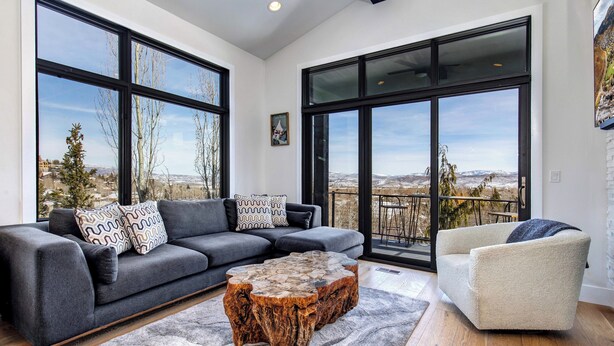 A modern living room with a grey sofa, a white armchair, and a unique wooden coffee table. Large windows offer a view of the outdoors. - Canyons Village Retreat with On-Demand Shuttle, Hot Tub by Park City Lodging (Park City)