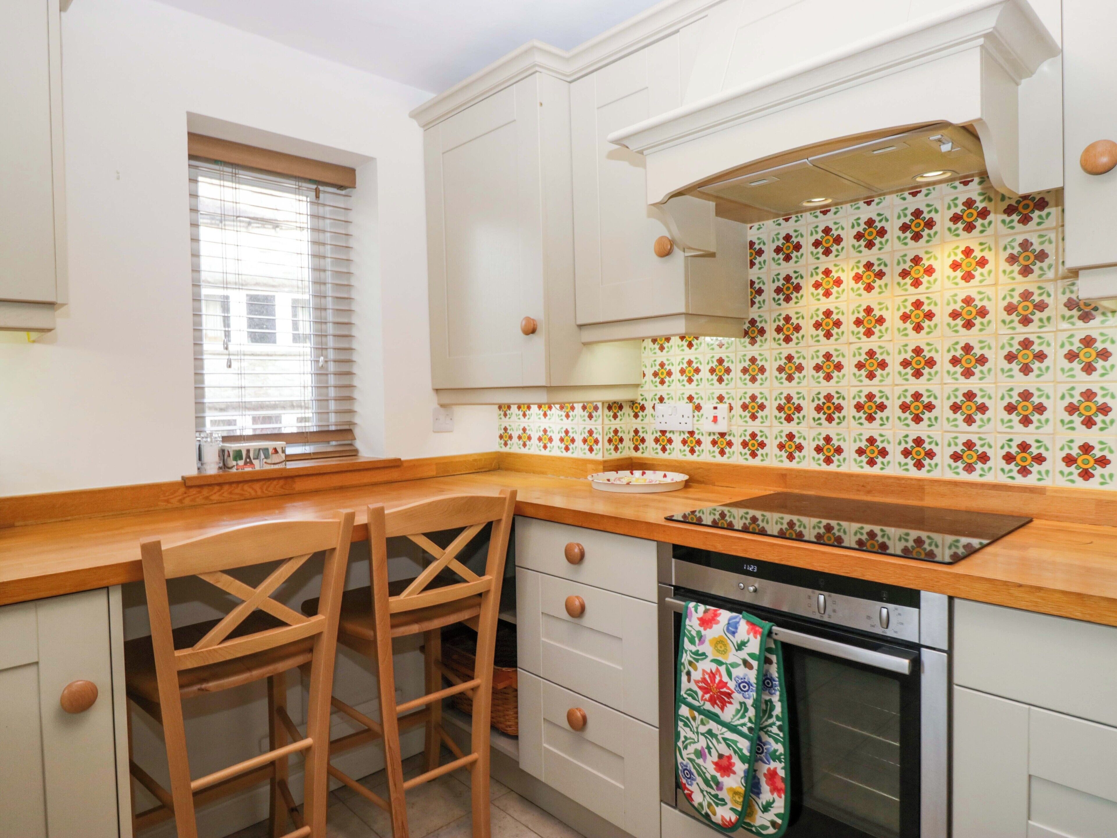 A kitchen with white cabinets, a wooden countertop, a window with blinds, and a colorful backsplash.