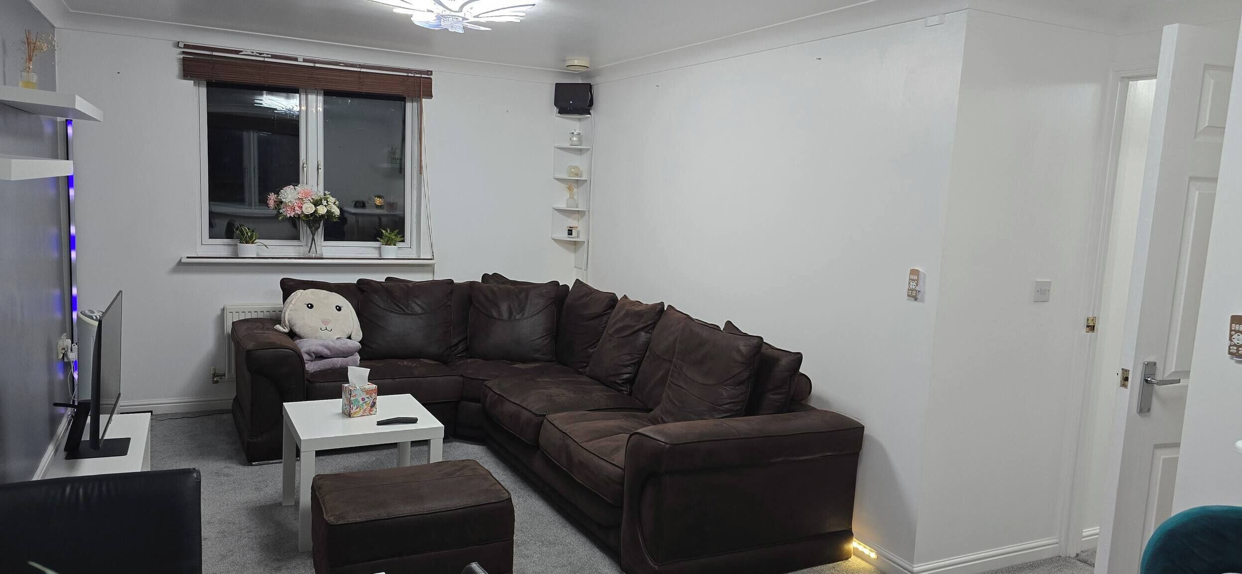 Una sala de estar moderna con un sofá de cuero marrón, una mesita de centro blanca y un pequeño reposapiés. Se ve una ventana con vistas al exterior, un radiador y una estantería con objetos decorativos.