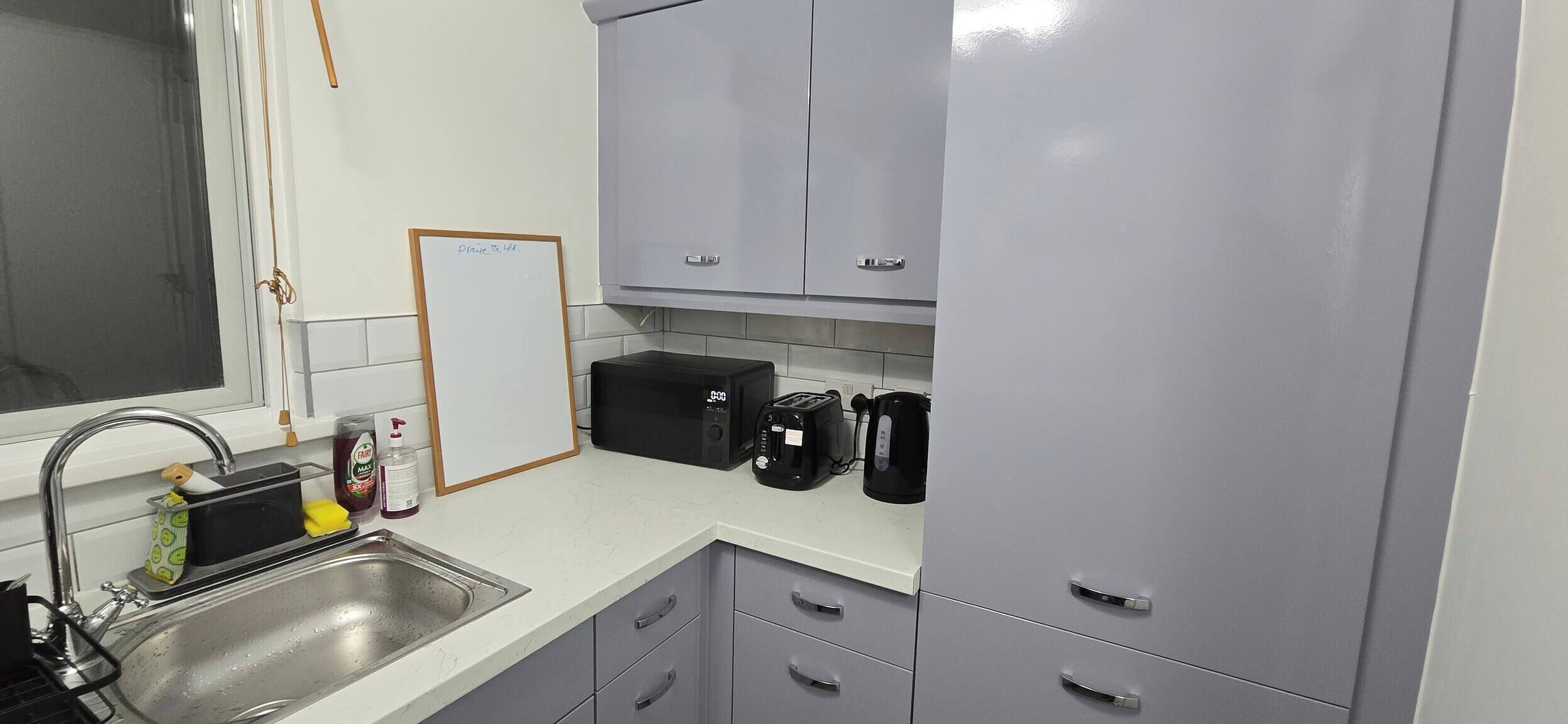 A compact kitchen with a sink, a microwave, a toaster, and a kettle. There is a whiteboard on the wall and a cabinet above the counter.