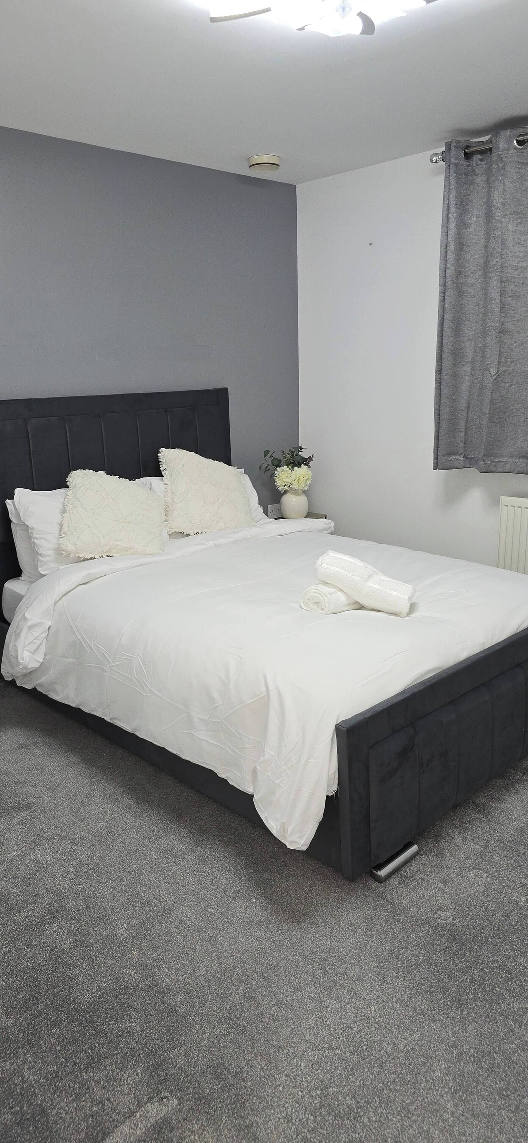 A neatly made bed with white linens and pillows, a grey headboard, and a grey carpeted floor.