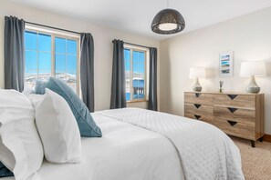 Une chambre à coucher avec un lit, deux fenêtres avec des rideaux, une commode avec des lampes et un luminaire suspendu.