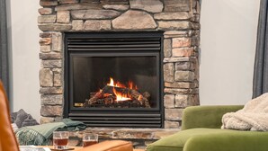 A stone fireplace with a black frame, a green armchair, and a wooden table with glasses and a blanket. - Teton Homestead: White Pine w/ Hot Tub & Views (Victor)