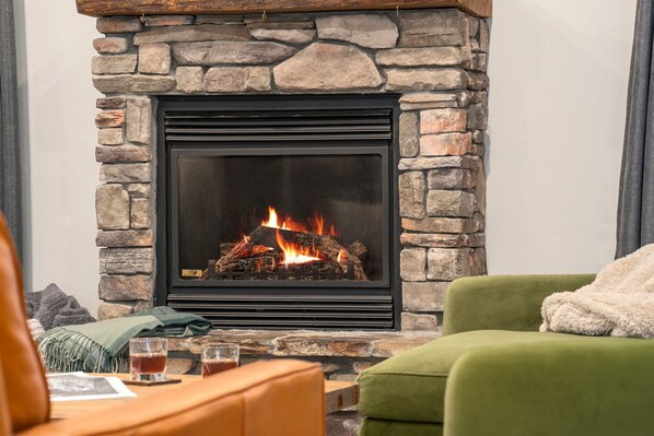A stone fireplace with a black frame, a green armchair, and a wooden table with glasses and a blanket. - Teton Homestead: White Pine w/ Hot Tub & Views (Victor)