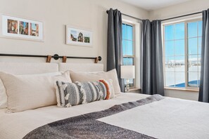 Een net opgemaakt bed met een hoofdeinde, meerdere kussens en een deken. Twee ramen met gordijnen, waarvan er een uitkijkt op een besneeerd landschap.
