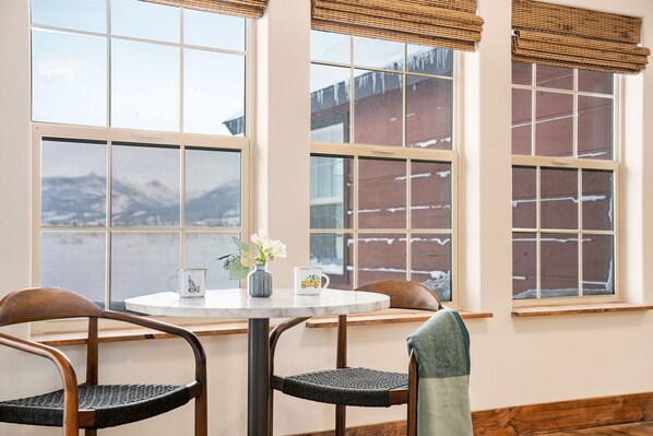 Un coin repas comprenant une table ronde, deux chaises et une vue sur un paysage enneigé, visible à travers de grandes fenêtres équipées de stores.