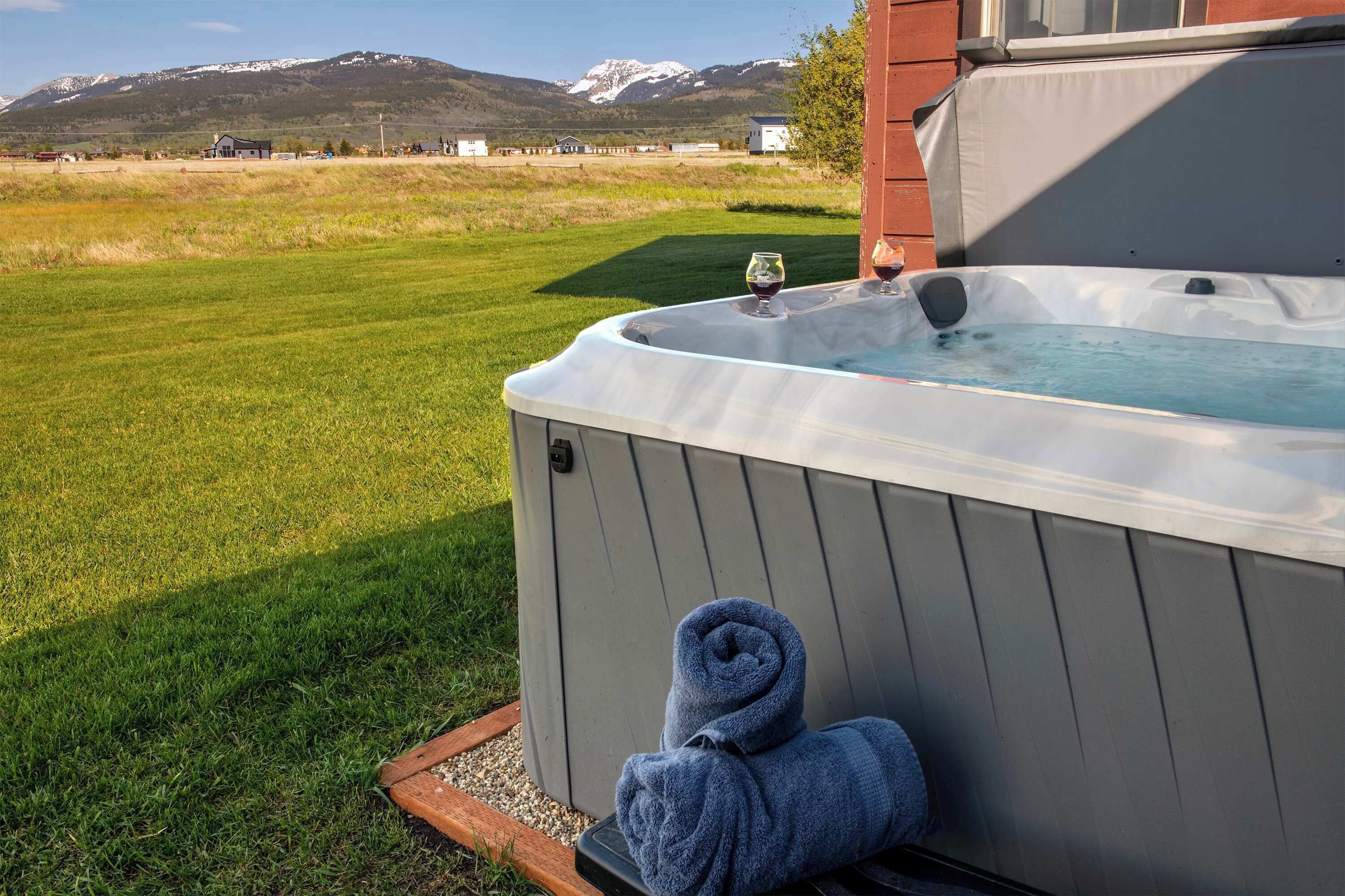 A hot tub with a towel and wine glass, set against a backdrop of a grassy field and mountains.