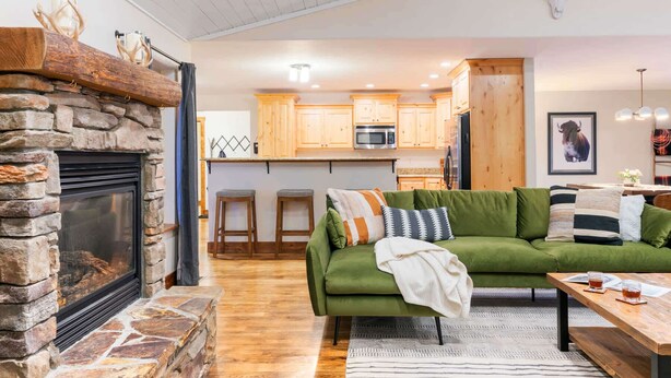 A cozy living room with a stone fireplace, a green sofa, and a kitchen in the background. - Teton Homestead: White Pine w/ Hot Tub & Views (Victor)