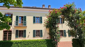 A two-story building with a balcony on the second floor, a garden with blooming flowers, and a clear blue sky. - Country House Amista by Italita (Nizza Monferrato AT)