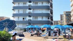 Een hotel met meerdere verdiepingen, met balkons, een zwembadgedeelte met ligbedden en parasols, en een zandstrand.
