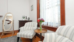 A room with two patterned armchairs, a round wooden table with a vase of flowers, a mirror, a dresser, and a window with curtains. - Charming Conshohocken B+B Near Philly (Conshohocken)