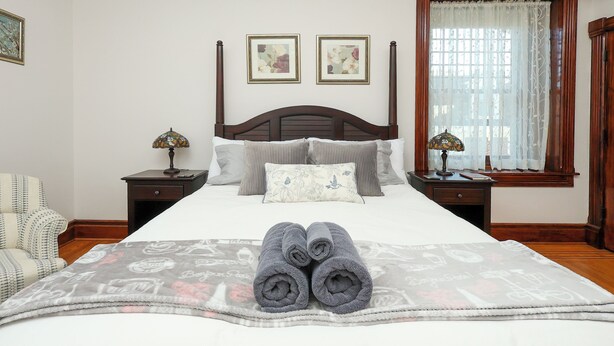 A neatly made bed with a patterned blanket, two bedside lamps, a chair, and a window with curtains. - Charming Conshohocken B+B Near Philly (Conshohocken)