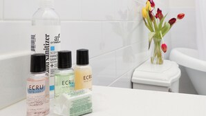 A bathroom with white tiles, a white toilet, and a sink with various toiletries including a bottle of ECRU shower gel, a hand sanitizer, and a bottle of ECRU body wash. - Charming Conshohocken B+B Near Philly (Conshohocken)