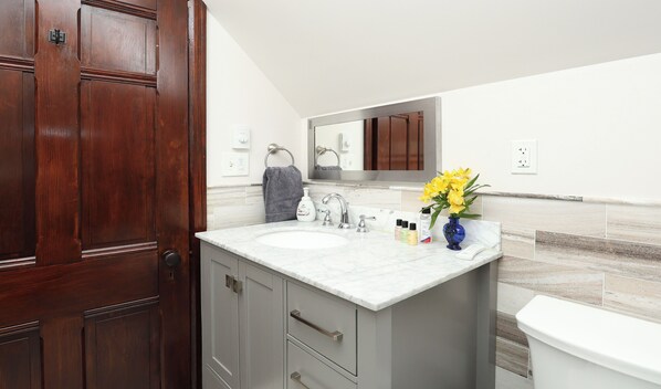 Un baño con puerta de madera, lavamanos de mármol, espejo, toallero, un jarrón con flores y un inodoro.