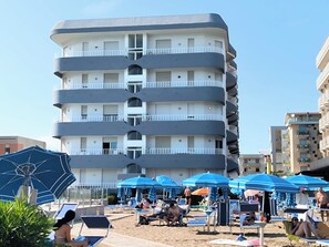 A multi-story hotel with balconies, a pool area with sun loungers and umbrellas, and a sandy beach. - Bright Holiday Home With Balcony (Jesolo)