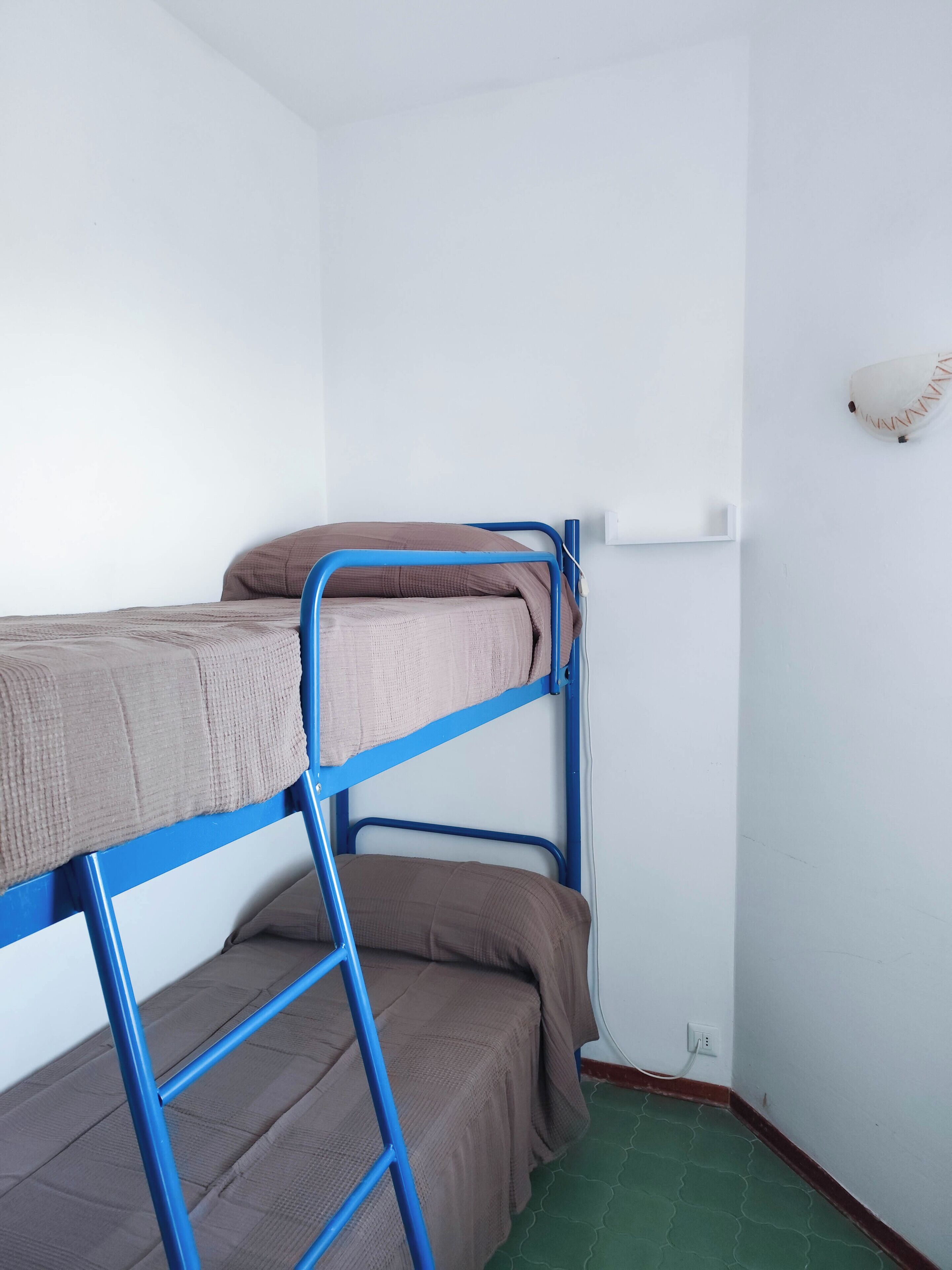 Una cama litera con barandillas de metal azul y ropa de cama marrón en una habitación con paredes blancas y suelo verde.