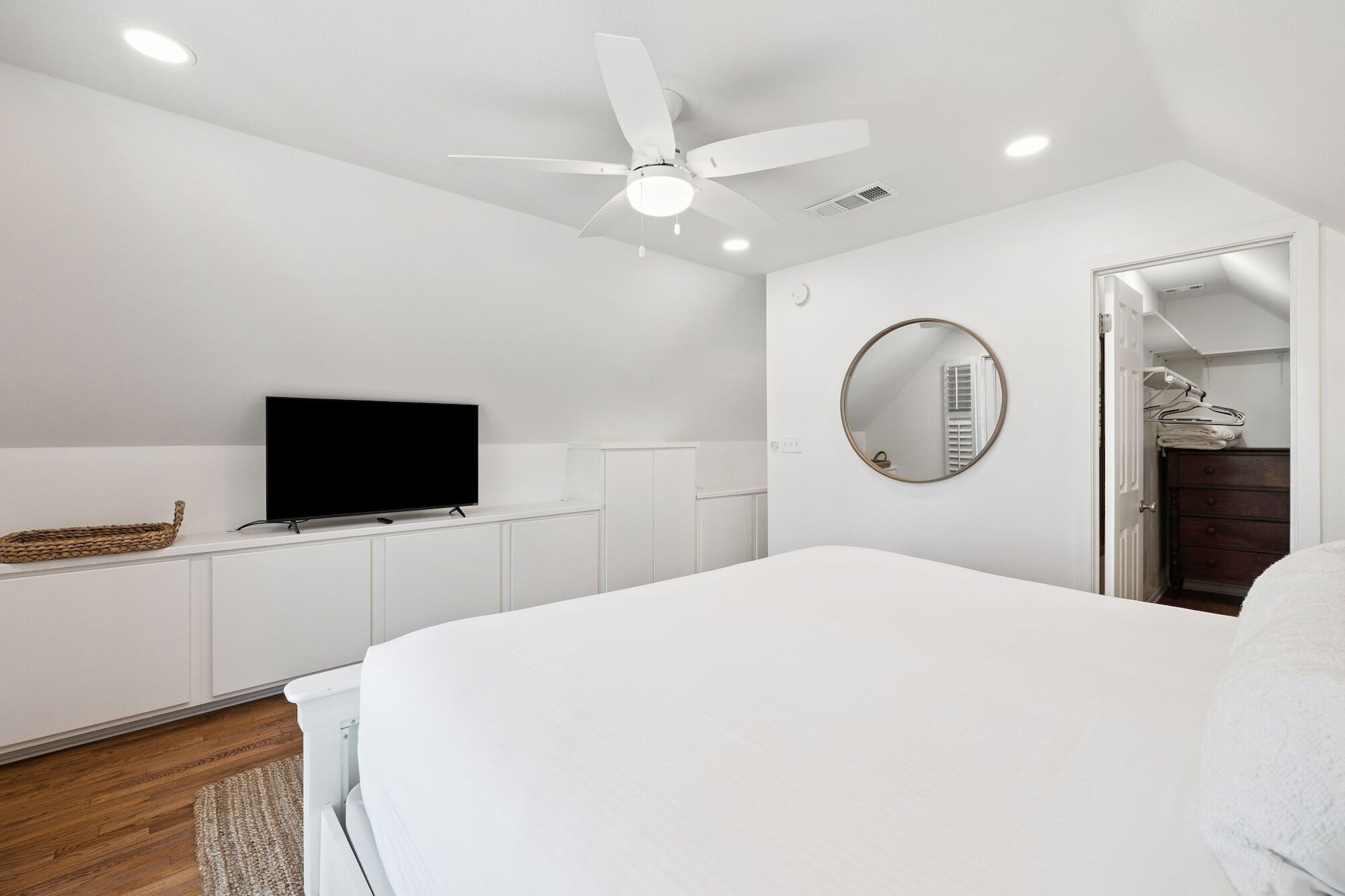 A bedroom with a bed, a TV on a white cabinet, a ceiling fan, and a round mirror on the wall.