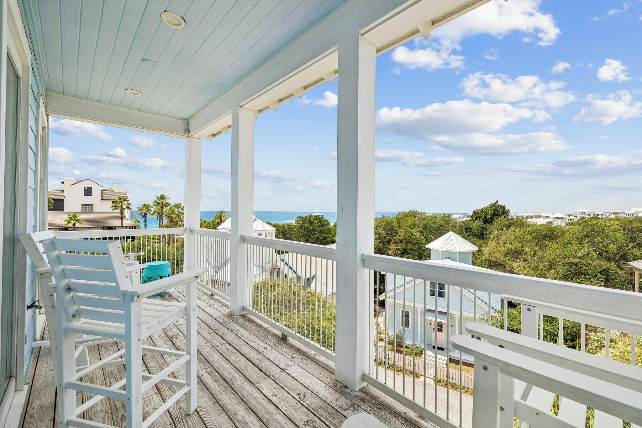A wooden deck with white railings, a blue chair, and a view of the ocean and buildings in the background.