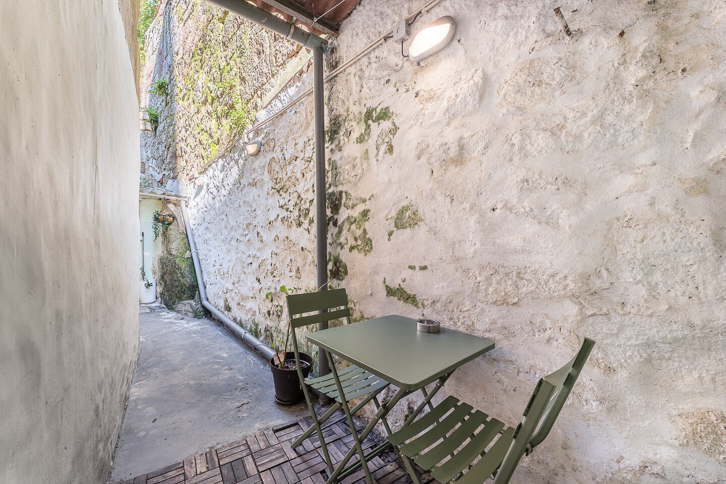 A small outdoor seating area with a table and chairs, a potted plant, and a light fixture on a textured wall.