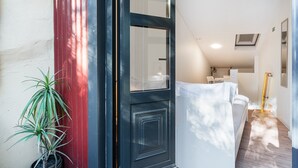 L’entrée d’un hôtel, avec une porte sombre, un pot de fleurs, un canapé blanc et un couloir menant à un espace cuisine.