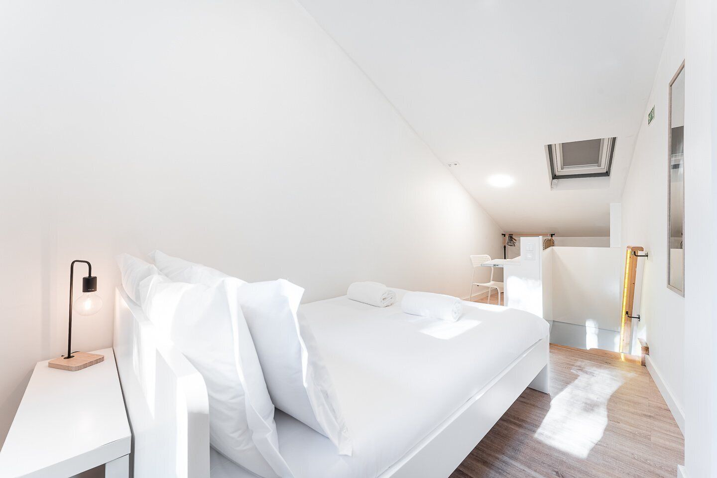 Une chambre d’hôtel moderne, décorée dans des tons de blanc, avec un lit soigneusement fait, une table de chevet avec une lampe et un petit bureau.