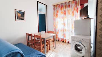 Quarto pequeno e limpo com cama, mesa e cadeiras, máquina de lavar roupa e janela com cortinas.
